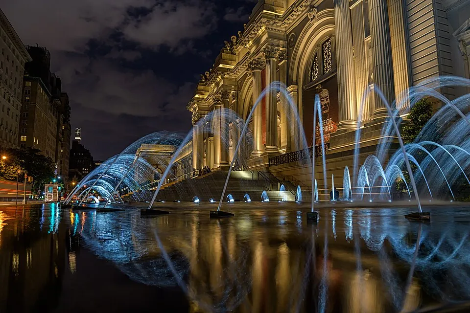 The Met's Fifth Avenue facade floodlit at night behind the plaza fountains